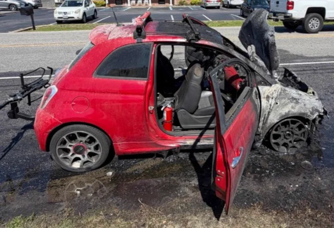 A small red car, severely damaged by fire, is parked on the side of the road. The front is charred, with the hood open and the interior partially exposed. A bike rack is mounted on the back. Surrounding the vehicle, the pavement is wet, likely from firefighting efforts. Other cars and a building are visible in the background.