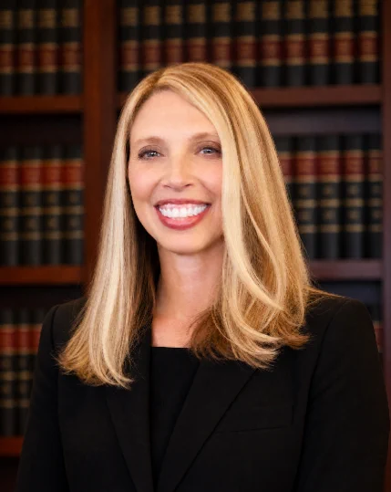 Professional headshot of a woman with long blonde hair wearing a black blazer and black top. She is smiling directly at the camera. The portrait is taken indoors with shelves of law books in soft focus behind her, creating a formal judicial or legal office setting.