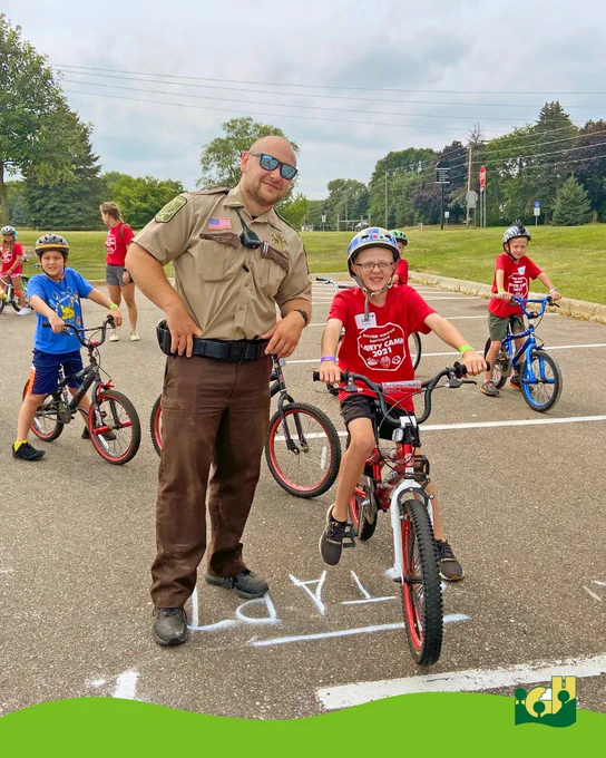 Children wearing helmets ride bikes in a parking lot while a uniformed officer stands nearby supervising the activity.