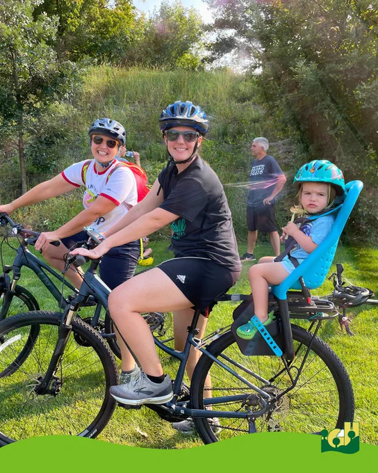 Two adults wearing helmets ride bicycles on a grassy path, with a child in a blue helmet seated in a rear bike seat on one of the bikes.
