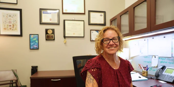 This image shows a person sitting in an office. The walls are adorned with multiple framed certificates and awards. The desk features an office phone, stationery, and papers. The person is smiling and wearing glasses.