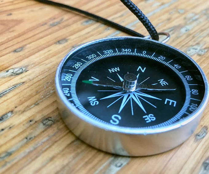 A silver-rimmed compass rests on a wooden surface, displaying cardinal directions with a needle pointing north. The compass features degree markings around its edge.