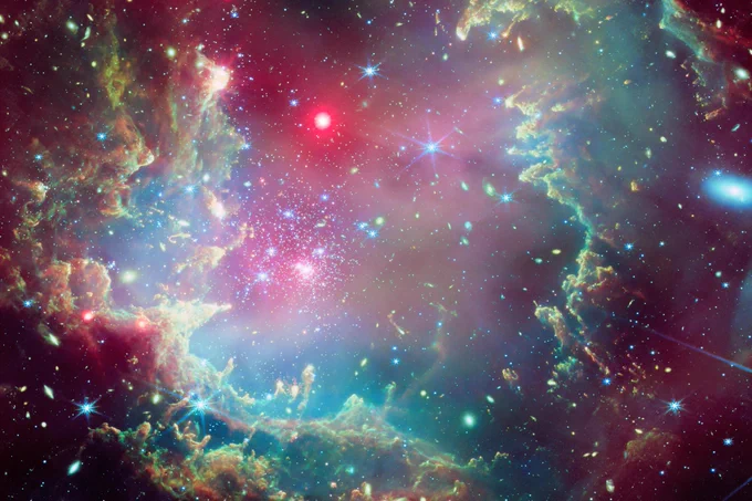 Imagen compuesta del cúmulo estelar NGC 602, apodado la “corona cósmica”, que combina datos del Observatorio de rayos X Chandra y el telescopio espacial James Webb de la NASA. La imagen, captada por Webb, muestra una llamativa formación parecida a un anillo de gas y polvo de colores (en naranja, amarillo, verde y azul) que se asemeja a una corona. En el centro y en toda la corona hay puntos rojos brillantes que representan las emisiones de rayos X detectadas por Chandra, y que muestran a estrellas masivas jóvenes y calientes que impulsan intensos vientos estelares. El cúmulo, situada en la Pequeña Nube de Magallanes, a unos 200.000 años luz de la Tierra, se observa en un contexto de innumerables estrellas y galaxias lejanas.  