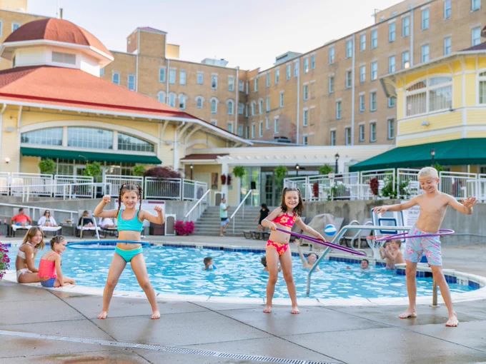Children having fun with hula hoops by French Lick Springs Hotel pools. The building in the background is a grand, multi-story structure with a red-roofed section. People are lounging around the pool, enjoying a sunny day.