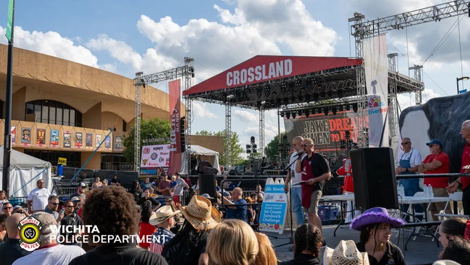 Outdoor event with a stage labeled "CROSSLAND," hosting live music and a performer speaking into a microphone. Attendees, some wearing hats, gather in front of the stage. The Wichita Police Department logo is visible in the foreground.