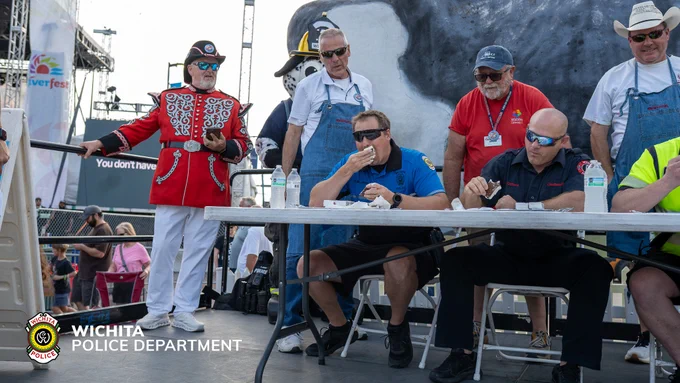 Four men are sitting and eating ice cream sandwiches; several individuals stand nearby, one applauding. A large cow model is visible, and the "Wichita Police Department" logo appears in the image.
