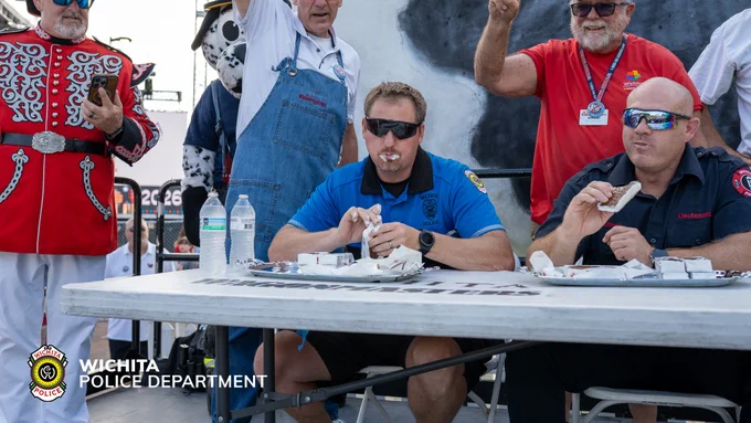 Four men are sitting and eating ice cream sandwiches; several individuals stand nearby, one applauding. A large cow model is visible, and the "Wichita Police Department" logo appears in the image.