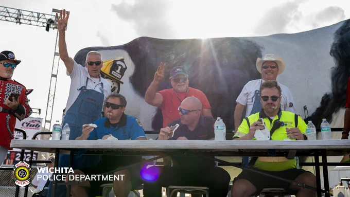 Four men are sitting and eating ice cream sandwiches; several individuals stand nearby, one applauding. A large cow model is visible, and the "Wichita Police Department" logo appears in the image.