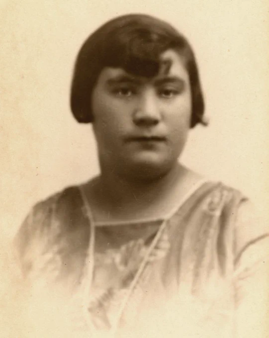 Sepia-toned portrait of a woman wearing a vintage blouse with delicate lace details at the neckline. The subject has a short, bobbed haircut and is looking directly at the camera with a serene expression.
