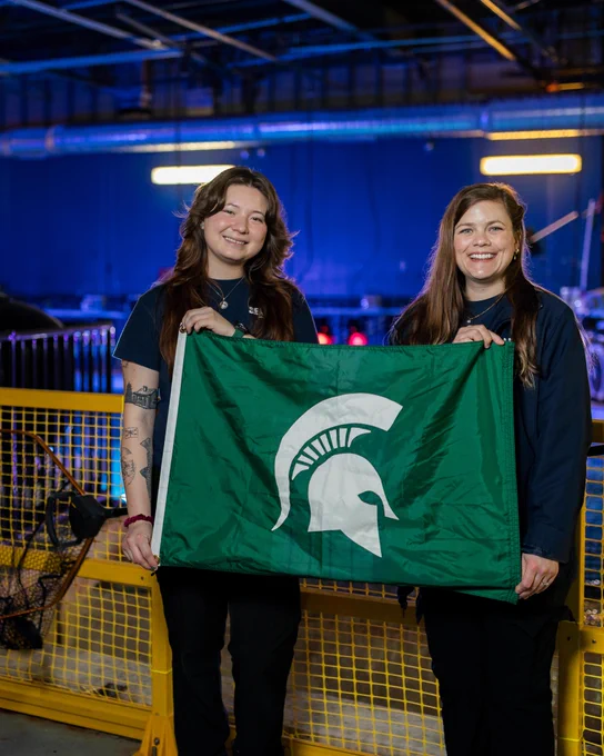 Two smiling people, one with tattoos, hold a green Michigan State University flag with a white Spartan helmet. They stand in an aquatic facility with blue lighting, a yellow safety barrier, and a fishing net visible to their left.