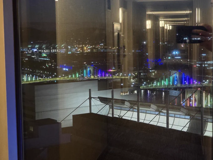 shot of bridges at night lit up in colors, through the window which also reflects the hotel hallway