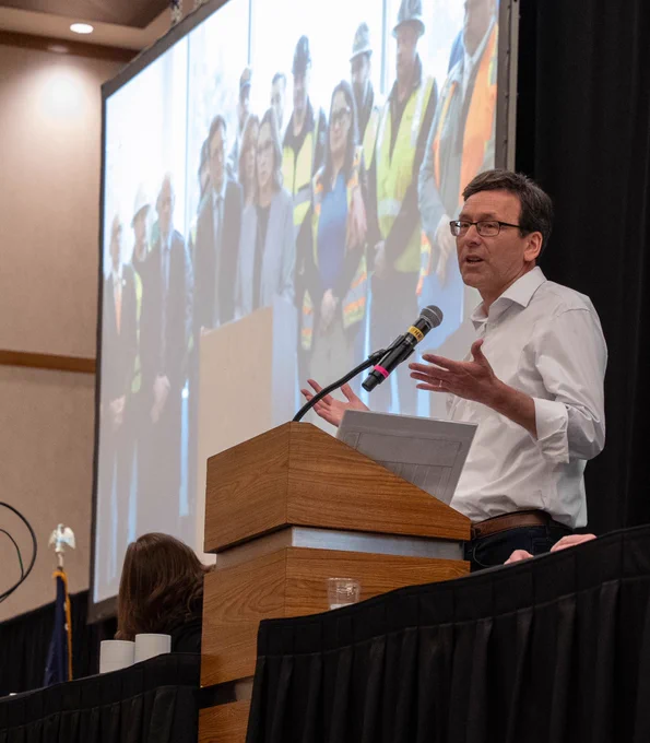 Bob speaking behind lectern; image on projector with individuals in safety vests from press conference on Interstate Bridge