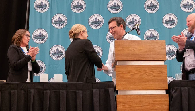 Bob shaking hand of individual on stage, 2 others applauding
