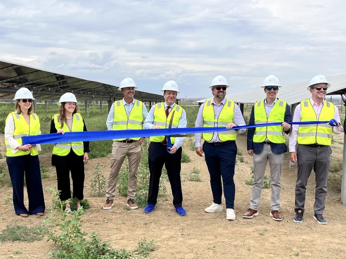 Group of seven people wearing safety vests and helmets holding a blue ribbon at a solar panel installation site.