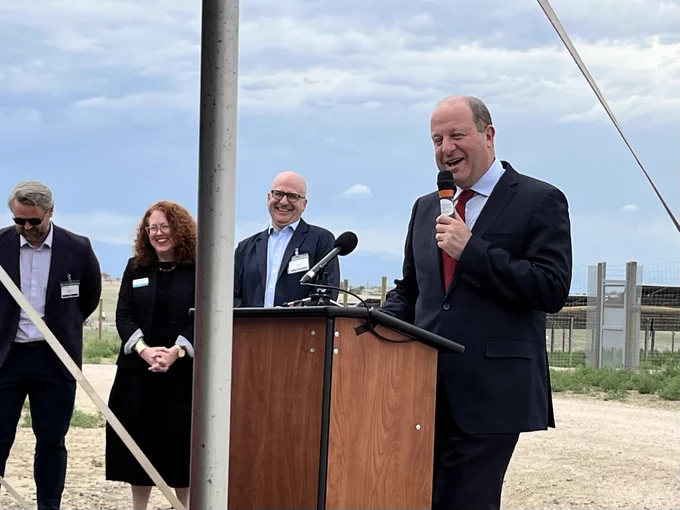 A man in a dark suit and red tie speaks into a microphone at a podium outdoors with three people standing behind him against a cloudy sky.