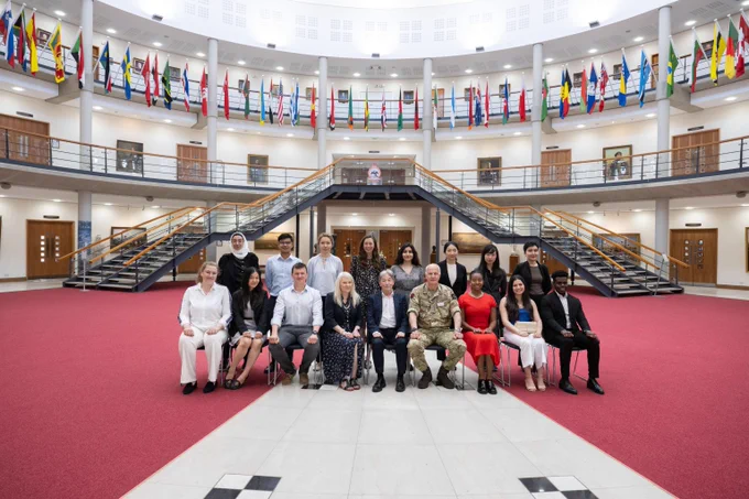 Group portrait of diverse professionals and military personnel seated and standing in a spacious hall with international flags overhead.