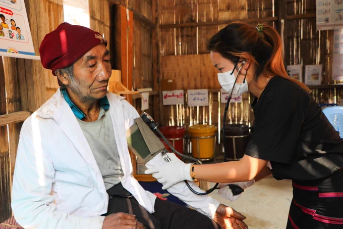 A young female doctor in a mask taking the blood pressure of an older man in a white coat and red hat. They are in a wooden building
