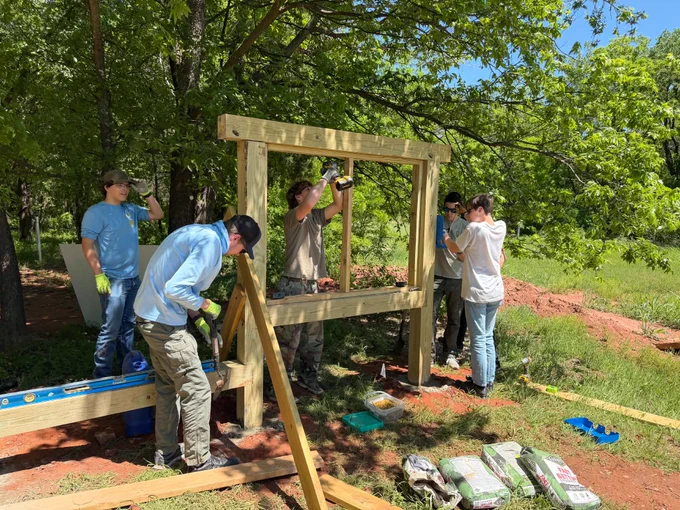 Several volunteers work together outdoors to construct a wooden structure in a wooded area. Lumber, tools and construction materials are scattered around the work site as participants assemble the frame under sunny conditions. Green trees and vegetation surround the project area.
