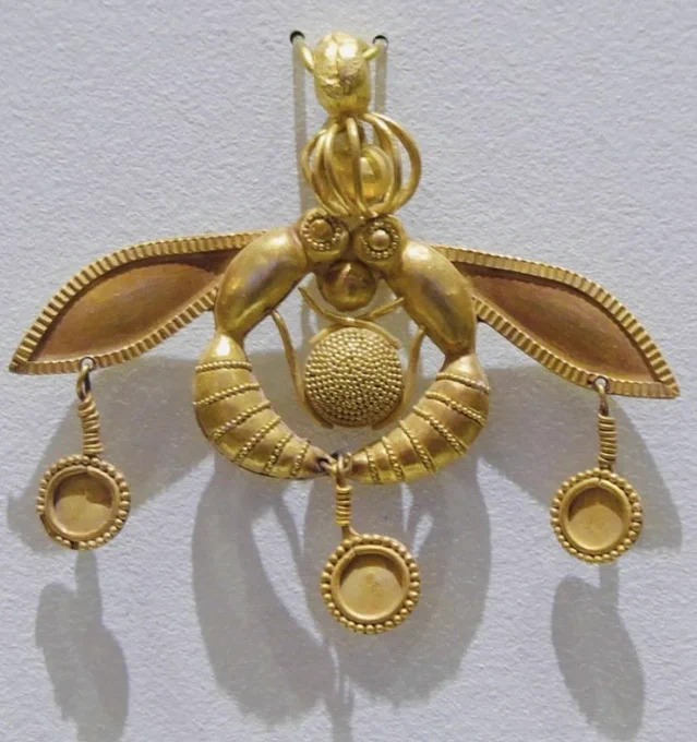 The image shows a circa 3,800 year-old Minoan gold pendant known as the ‘Malia Bee Pendant’. It is said to represent two bees facing each other in the process of depositing a drop of honey in their honeycomb. Their heads and the ends of their bodies are joined. Each insect has a single wing behind the body. In the centre of the pendant, framed by the curving bodies, the bees hold a round, granulated honeycomb between their legs, and a round 'drop' of honey balanced between their mouths. On their heads is a spherical filigree cage enclosing a solid gold sphere. Hanging down are three circular gold discs decorated with beaded border; a disc from each wing and one from where their end of their bodies meet at the sting. The pendant measures 4.6 cm maximum diameter. Excavated from the Minoan Bronze Age cemetery of Chrysolakkos at ancient Malia, Crete. Dated 1800 BC.