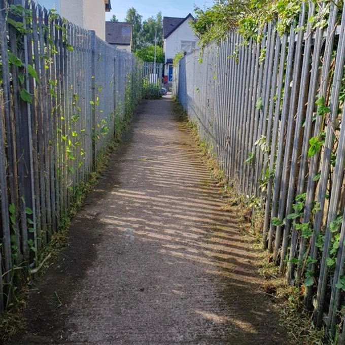 A narrow path between two tall metal fences with sunlight casting striped shadows.