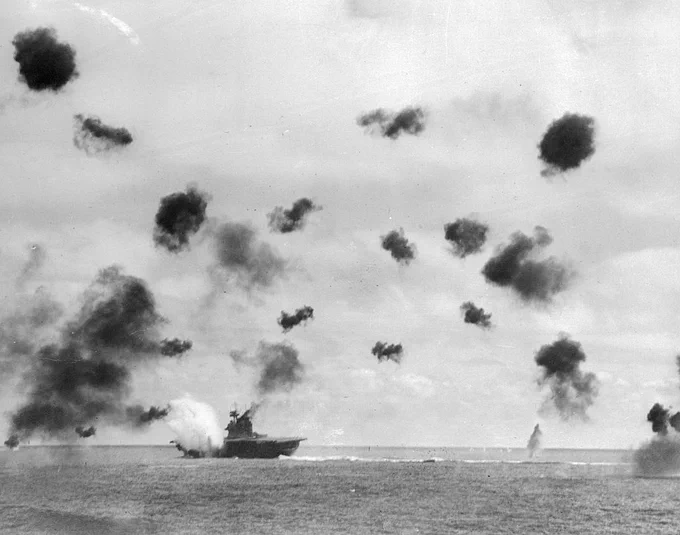 A historical photograph shows the USS Enterprise aircraft carrier at sea amid bursts of anti-aircraft fire during World War II. The sky is dotted with dark explosions, and the ocean is calm beneath the action.