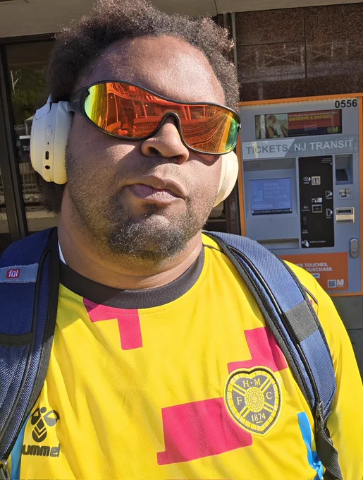 A selfie of myself outside. I am wearing sunglasses and headphones. The Hearts FC logo is featured on my top in the photo. Behind me is a ticket kiosk for NJ Transit