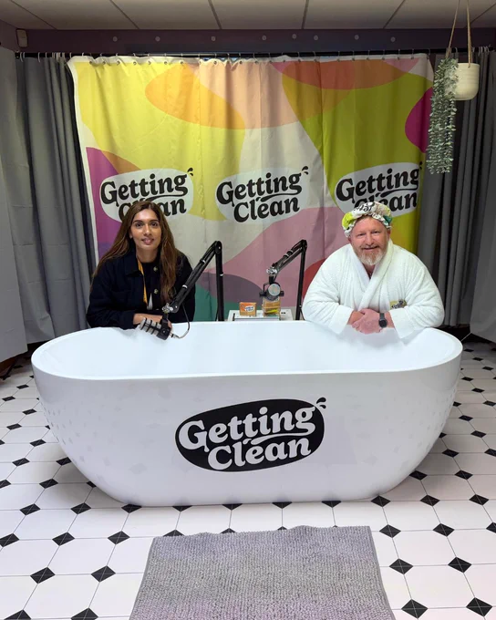 Barca-Leeds staff member poses by a Getting Clean bathtub with the Getting Clean founder. A Getting Clean branded shower curtain sits behind them, with a bathmat in the forefront.