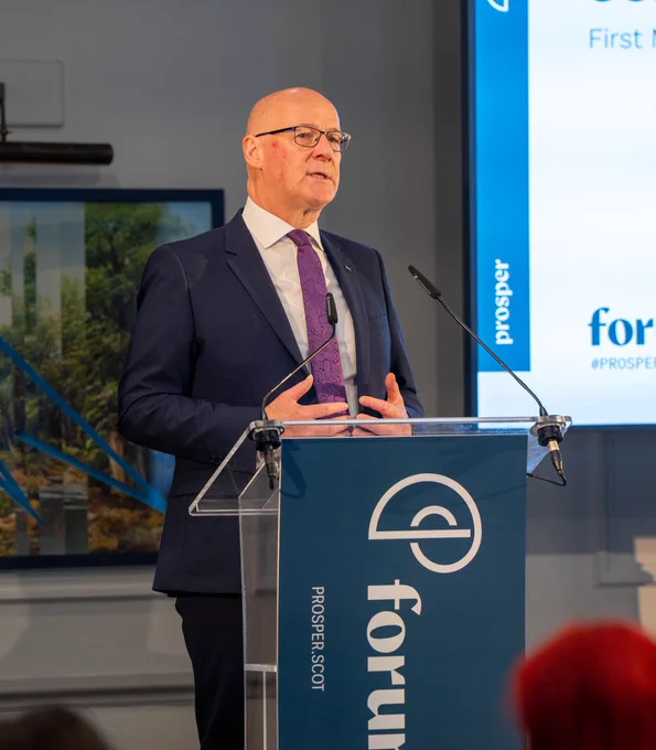 First Minister John Swinney standing at a lectern addressing people at the Prosper Forum. 