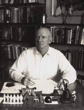 Colonel Russell P. "Red" Reeder Jr. sits at his desk in a white shirt, pen in hand over a sheet of paper, with a wall of books behind him and small toy infantry figures lined up on the desk in front of him. Black and white photo.