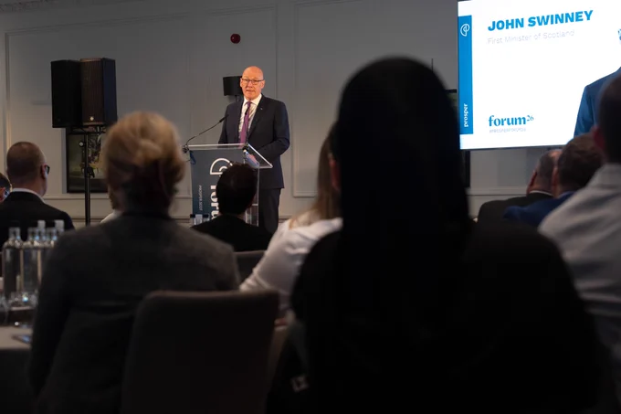 Photo of John Swinney speaking at a lectern at the Prosper Annual Forum event, with audience silhouettes in the foreground