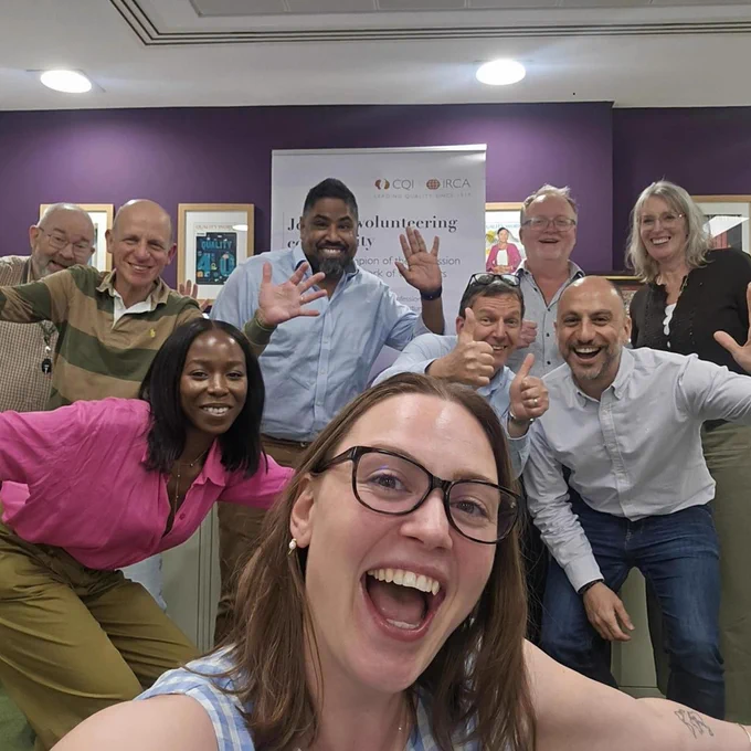London Network volunteers meet up at our office: a group of seven adults posing energetically in a room with purple walls and framed pictures, some giving thumbs-up and waving.