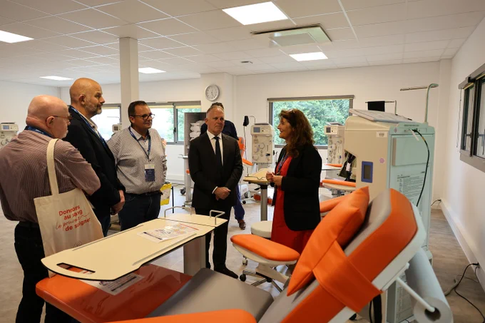 Cinq personnes, dont trois hommes en costume ou chemise et une femme en robe rouge et veste noire, discutent dans un centre médical moderne dédié au don du sang. Ils sont entourés de fauteuils oranges, de machines de prélèvement et d’appareils médicaux. L’un des hommes porte un sac avec l’inscription « Donnons notre sang, le pouvoir de soigner ». La scène se déroule dans une pièce lumineuse aux grandes fenêtres et à l’ambiance professionnelle.
