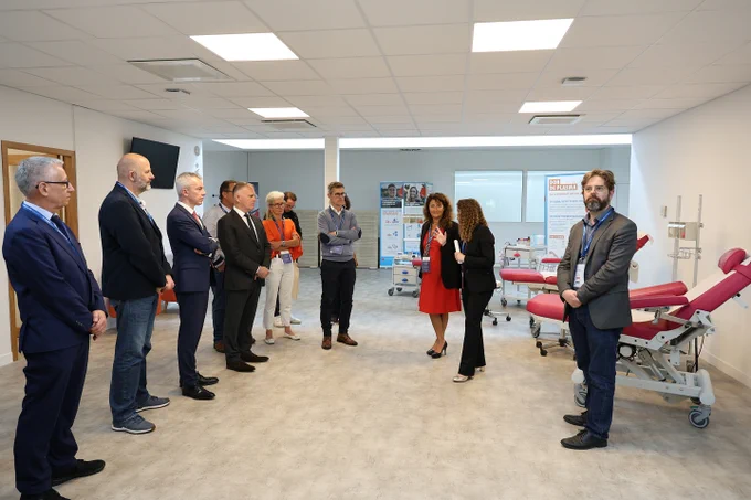 Un groupe de dix personnes, hommes et femmes en costume ou tenue professionnelle, se tient debout dans une salle moderne et lumineuse, équipée de fauteuils rouges pour prélèvements sanguins. Deux femmes échangent au centre du groupe. Des affiches sur le don de plasma sont visibles à l’arrière-plan. L’atmosphère est professionnelle, comme lors d’une visite officielle ou d’une inauguration dans un centre médical ou de don du sang.