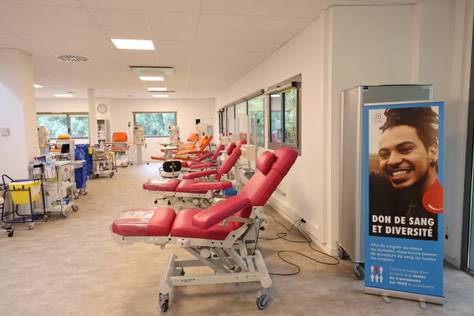 Salle de don du sang moderne, lumineuse et propre, avec plusieurs fauteuils de prélèvement rouges alignés le long d'une grande baie vitrée. À droite de l'image, un présentoir affiche une campagne sur le don de sang et la diversité, montrant le visage souriant d'un homme et un message incitant les personnes de toutes origines à donner leur sang. Plusieurs chariots médicaux sont visibles au fond de la pièce.