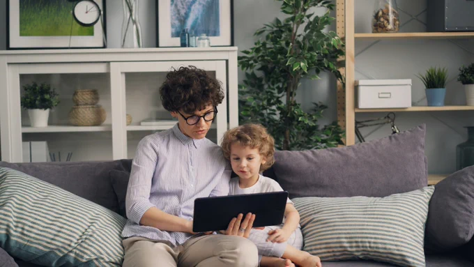 A woman sitting on a couch with a child next to her looking at an electronic device.