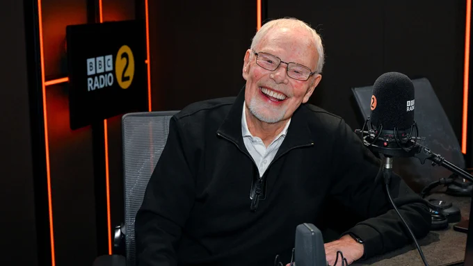 A man in a dark zip neck top and white shirt smiles to camera, laughing whilst sitting behind a desk in the BBC Radio 2 studio