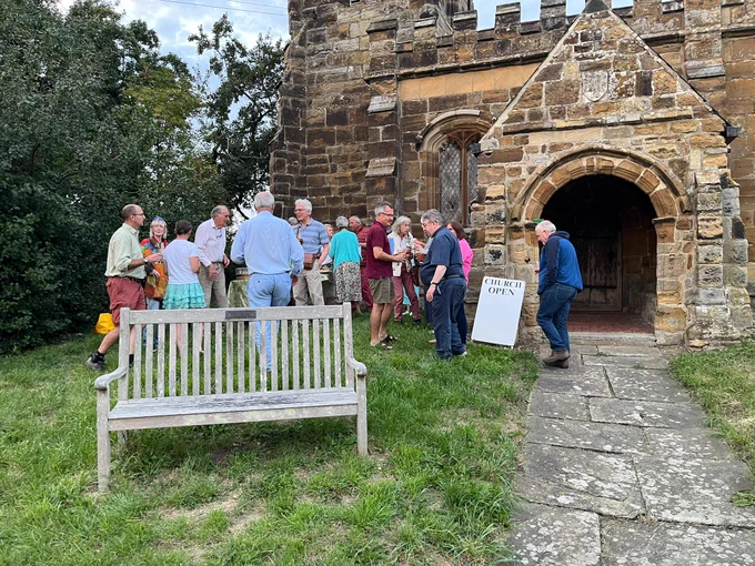 A group of people gathered in conversation outside a historic church. An A-Board displays the text "church open" in black on white ground. It is summer.
