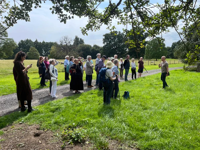 A group of people standing on a sunny country path looking up at a church, which is out of frame. A volunteer at the front addresses the group, giving a guided tour.