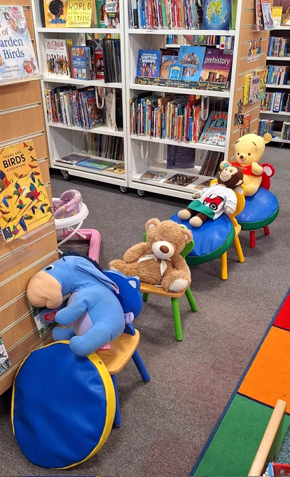 Eeyore, brown bear, monkey and Winnie the Pooh sitting on a line of chairs. Eeyore is slumped against the bookshelves