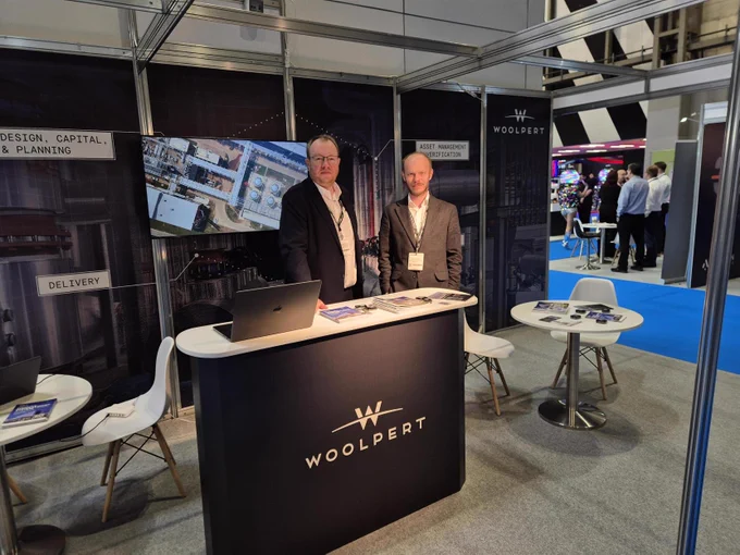 Matthieu Marsault and Adam Grewenda behind a Woolpert-branded booth at a Smart Manufacturing Week with promotional materials and an event showreel in the background.