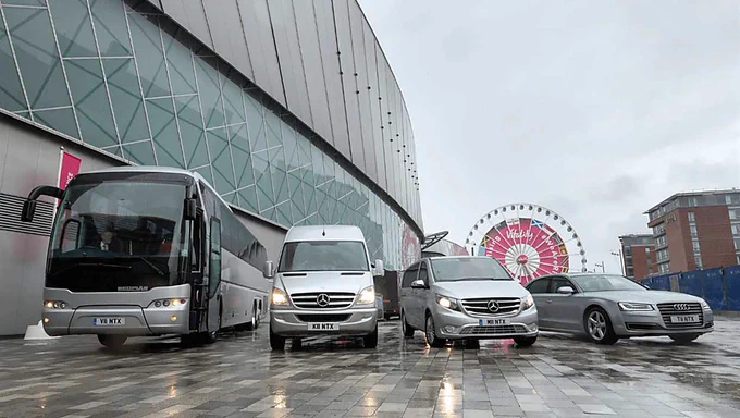 Four vehicles, a large coach bus, a high-roof van, a minivan, and a sedan, are lined up on a wet paved area outside a modern building with a glass façade. In the background, a lit Ferris wheel is visible. The vehicles display branded license plates, and reflections from the damp ground and overcast sky create a cool, muted atmosphere.