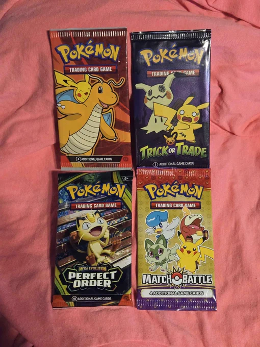 Four sealed Pokémon Trading Card Game packs arranged on a pink fabric background. The packs shown are a red 2024 McDonald’s Dragonite promo pack, a black 2023 Trick or Trade pack with Pikachu and Mimikyu, a Mega Evolution Perfect Order booster pack with Meowth artwork, and a 2023 McDonald’s Match Battle promo pack featuring Pikachu, Fuecoco, Quaxly, and Sprigatito.