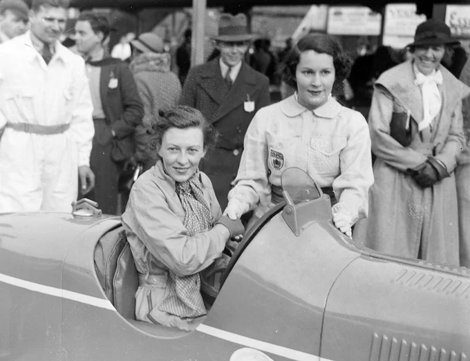 In the centre foreground watched on by observers in the pit area, Doreen Evans sits in her single seater MG, wearing race overalls and a long spotted scarf. Standing beside the car, wearing light coloured, BRDC badged overalls and a wide support belt, is Kay Petre - the who are shaking hands for the camera. 1935 BARC Opening Meeting, Brooklands.