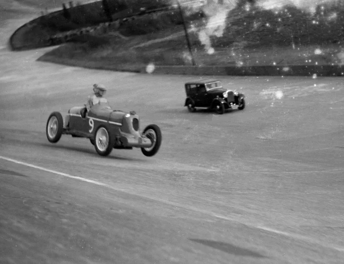 Shot from the top of the banking looking down at the track, Doreen Evans is partly airborne in her MG Midget Q Type after passing over a bump. Evans sits quite high in the cockpit of the single seater, open wheeled car. Further in the background on the flat part of the track, an unidentified road car can be seen. 1935 BARC Opening Meeting, Brooklands.