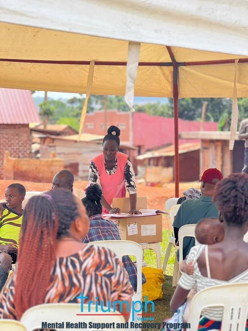 ​A view from behind the audience under a large outdoor tent looking toward a community health coordinator in a pink apron. She stands at a table resting on a cardboard box, organizing registration forms and files. Community members, including a mother holding her baby, sit in white plastic chairs listening attentively.