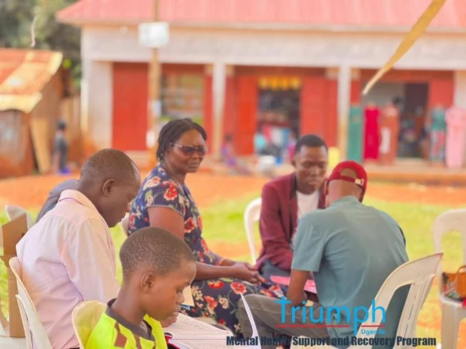 ​A close-up view under an outdoor tent showing one-on-one health counseling and registration sessions. In the foreground, a young boy looks down at papers, while behind him, a woman in a floral dress talks with a health team member. To the right, another peer educator in a teal shirt and red cap sits facing a young man, discussing outreach materials.