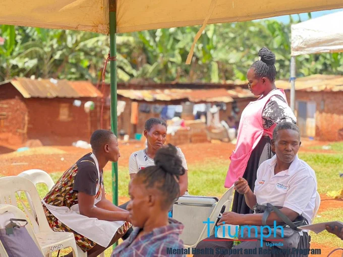 ​A community health worker wearing a pink apron stands under an outdoor tent, engaging with a group of women seated in white plastic chairs. One woman in the foreground holds a baby. The background shows red dirt ground and local storefronts, capturing an active and supportive community counseling atmosphere during the health outreach.