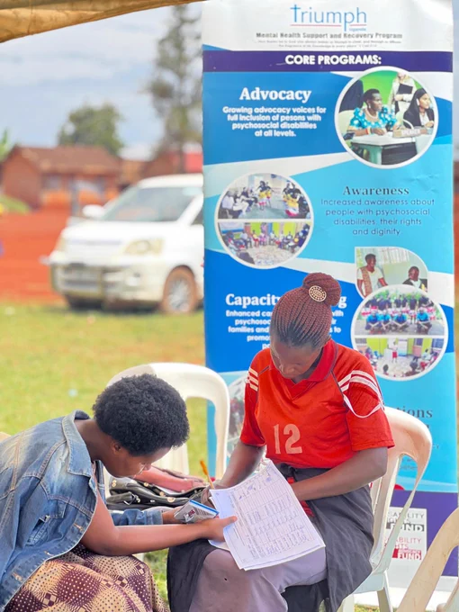 A young woman wearing a red jersey with the number 12 sits at an outdoor desk, meticulously filling out a registration data sheet. Next to her, an adolescent girl in a denim jacket leans over the table to assist or look at the form. Behind them stands a tall blue TRIUMPH banner detailing core programs like Advocacy, Awareness, and Capacity Building for full inclusion of persons with psychosocial disabilities.