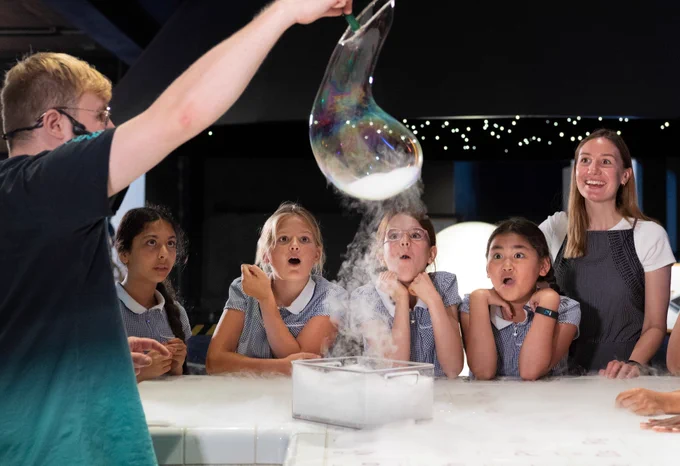 A group of young students in school uniforms observe a science experiment involving a large, colorful soap bubble. A Science Museum Explainer is demonstrating the experiment.