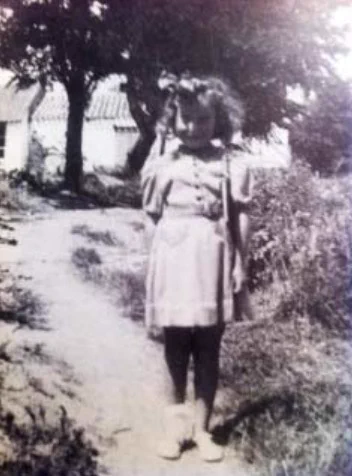 A vintage black and white photograph of a young girl standing on a dirt path with their hands behind their back. In the background, there are some small houses surrounded by trees. The child is wearing a dress and shoes, and has a bow in their hair.
